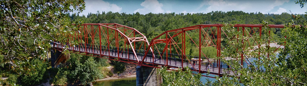 wide view of a red bridge spanning over a river framed by large, green trees 