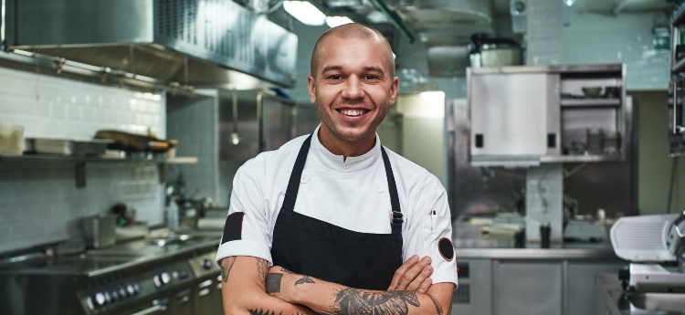 Bald man in commercial kitchen wearing black apron smiles at camera